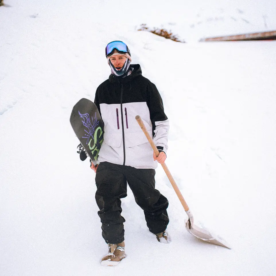 Jeune homme tenant son surf et une pelle à neige sur une piste de ski